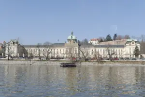 Blick über die Moldau auf die Straka-Akademie in Prag, ein großes historisches Gebäude mit hellen Fassaden und Türmen am Flussufer.