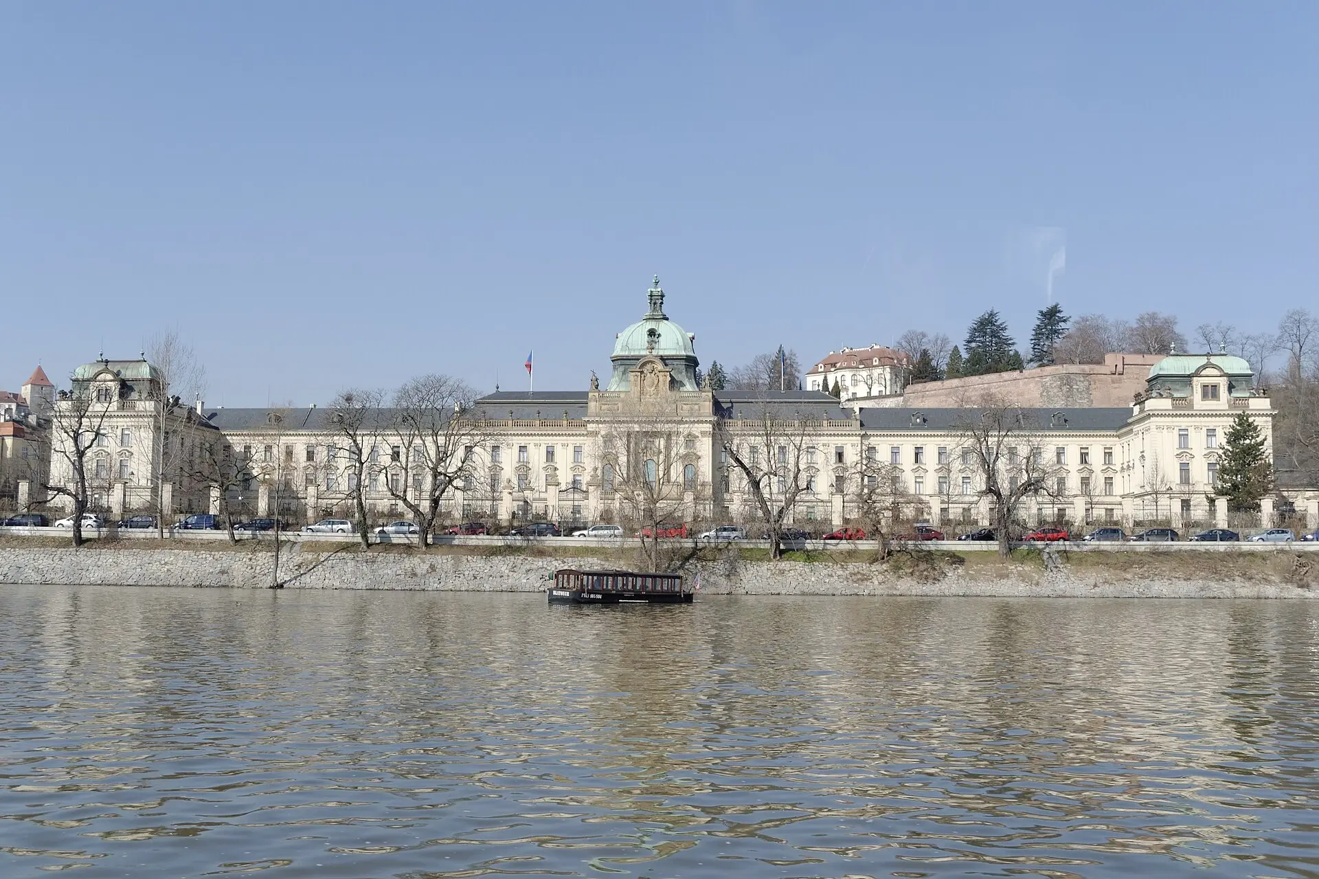 Blick über die Moldau auf die Straka-Akademie in Prag, ein großes historisches Gebäude mit hellen Fassaden und Türmen am Flussufer.