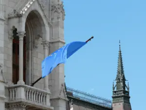 Blick auf das ungarische Parlamentsgebäude in Budapest mit einer wehenden EU‑Flagge im Vordergrund.